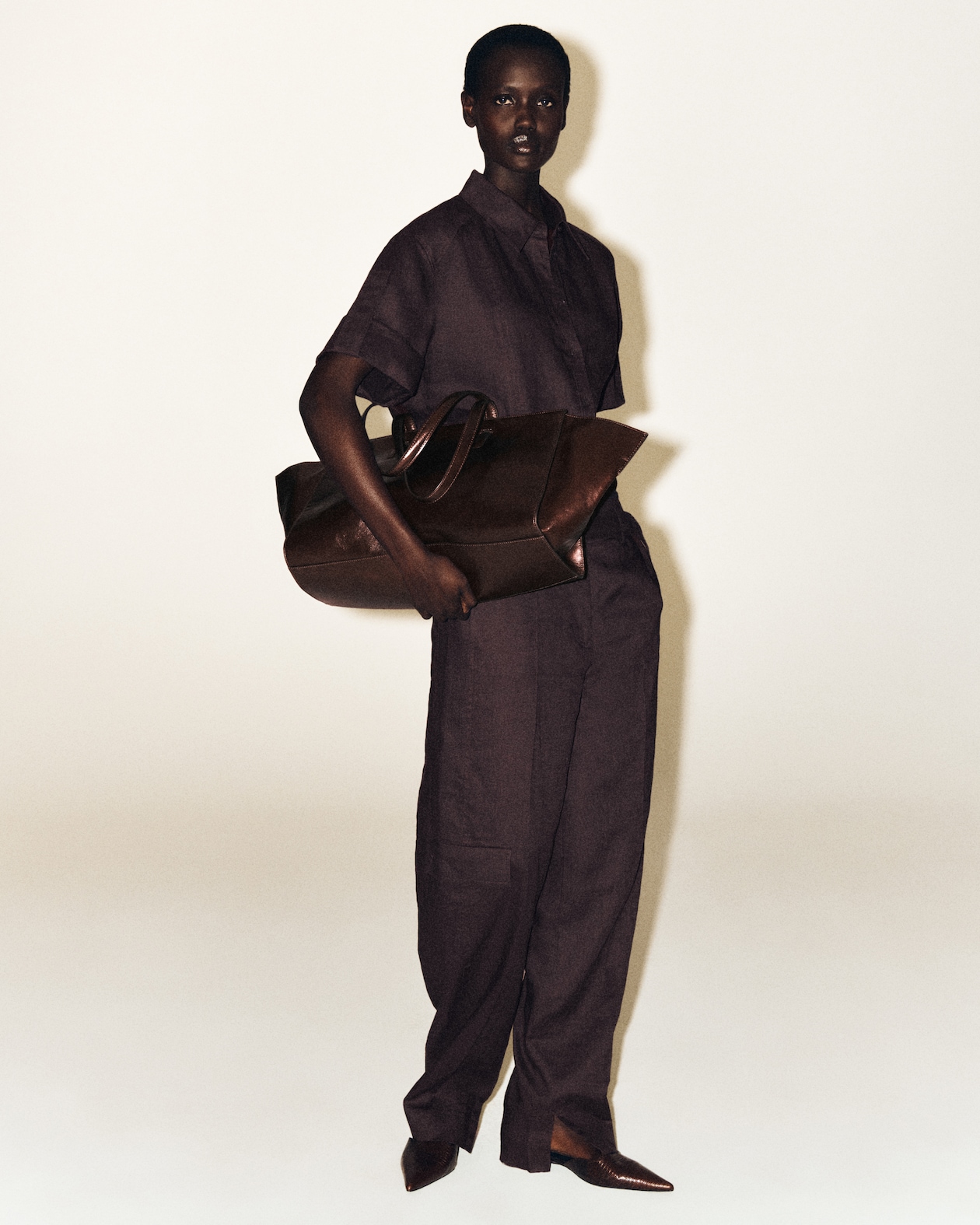 Person in a dark brown outfit holds a large brown leather bag, standing against a plain background.