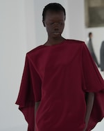 Placeholder for: A person wearing a flowing burgundy dress stands in a softly-lit room with blurred figures in the background.