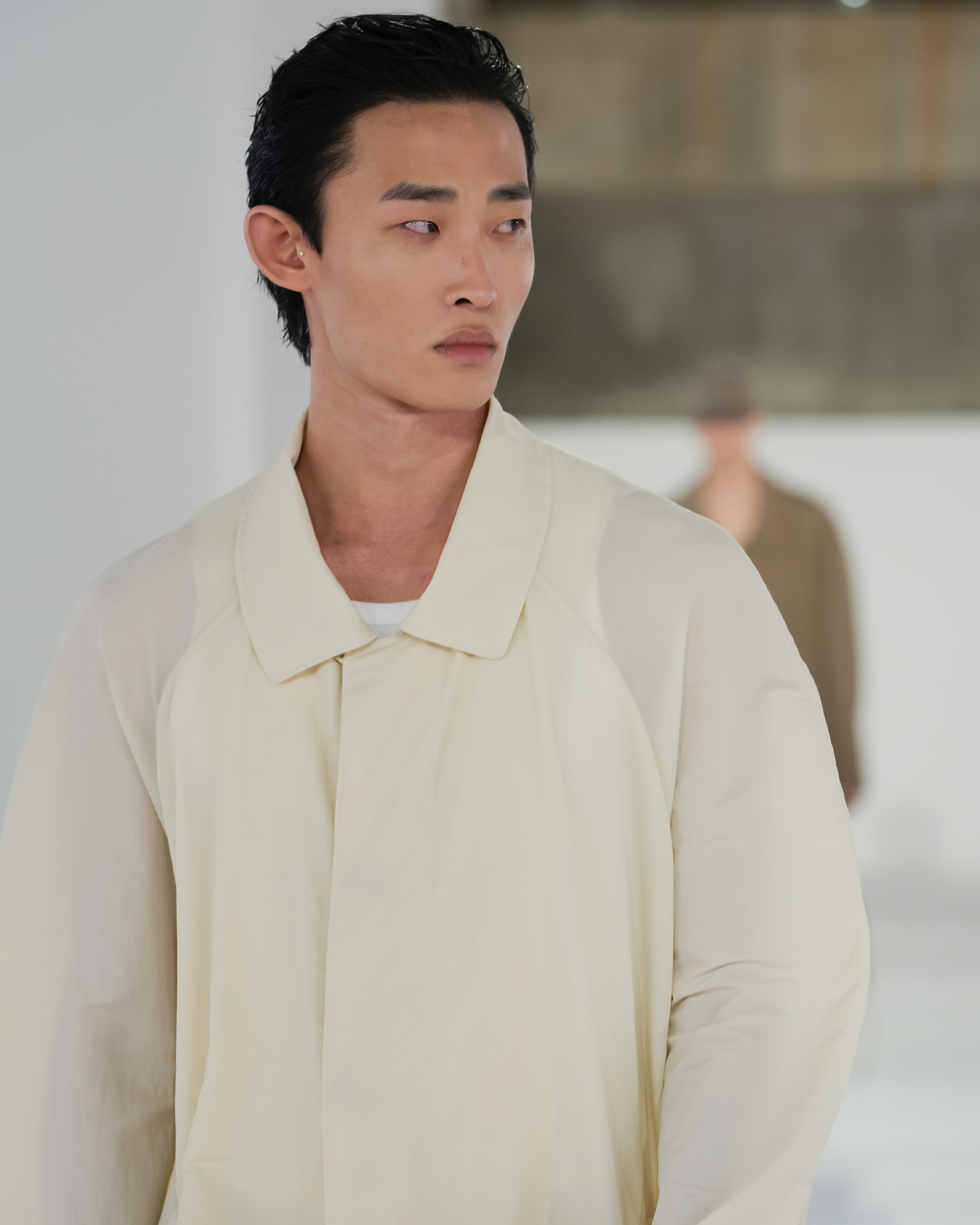 Person wearing a cream-colored coat, looking to the side, against a blurred background with another figure in soft focus.