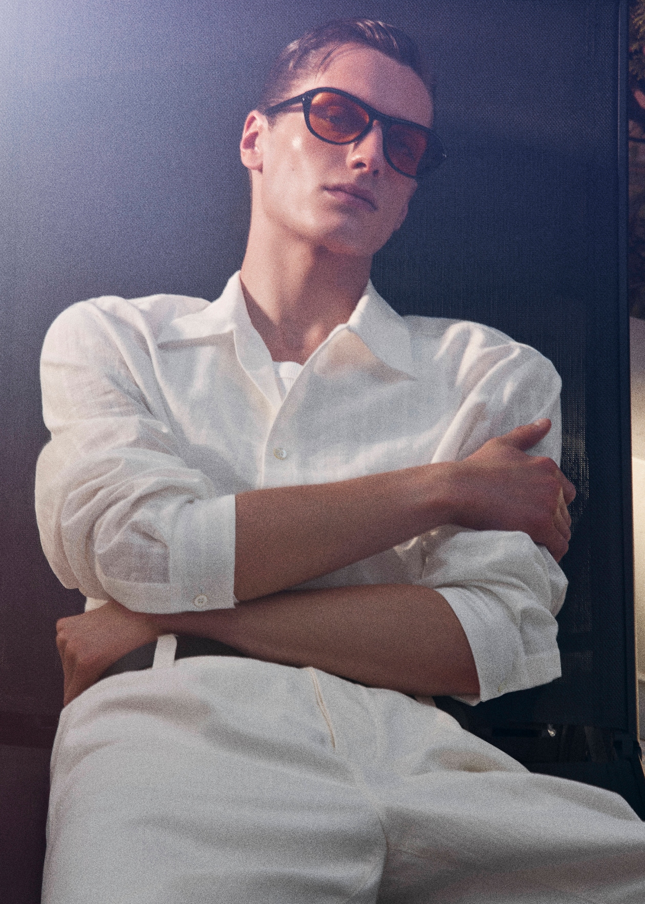 Person in a white shirt and pants with sunglasses, seated and leaning back with arms crossed, in a relaxed pose.
