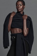 Placeholder for: A person poses confidently, wearing a black sweater and pants, with a belt at the waist. The background is a plain gray.