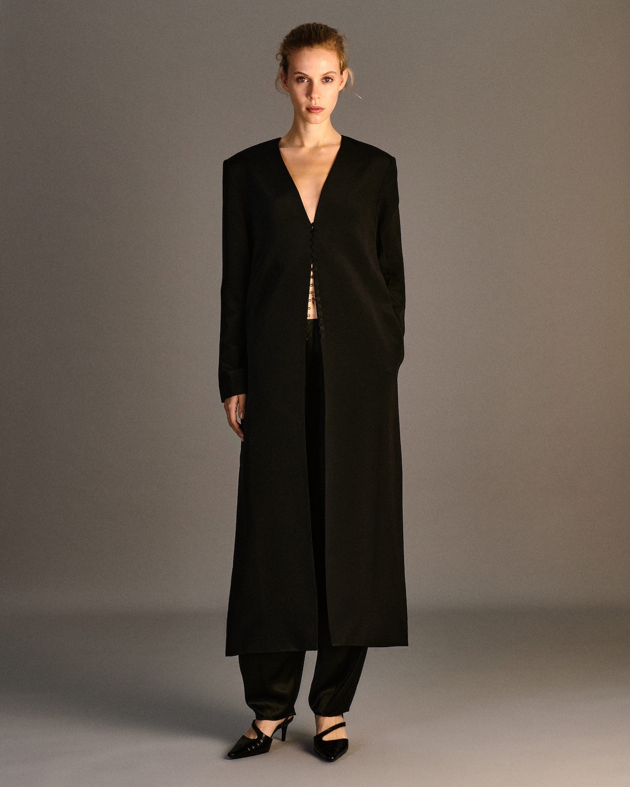A person wearing a long black coat with a deep V-neck, black pants, and black pointed heels. The background is a neutral gray.