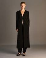 Placeholder for: A person wearing a long black coat with a deep V-neck, black pants, and black pointed heels. The background is a neutral gray.