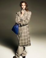 Placeholder for: A person in a plaid coat and beige trousers holds a blue bag, posing with a confident expression against a neutral background.