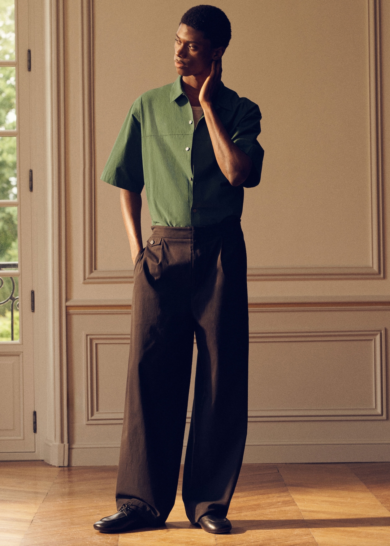A person in a green short-sleeved shirt and wide black pants stands in a sunlit room, touching their neck and looking to the side.