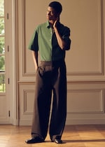 Placeholder for: A person in a green short-sleeved shirt and wide black pants stands in a sunlit room, touching their neck and looking to the side.