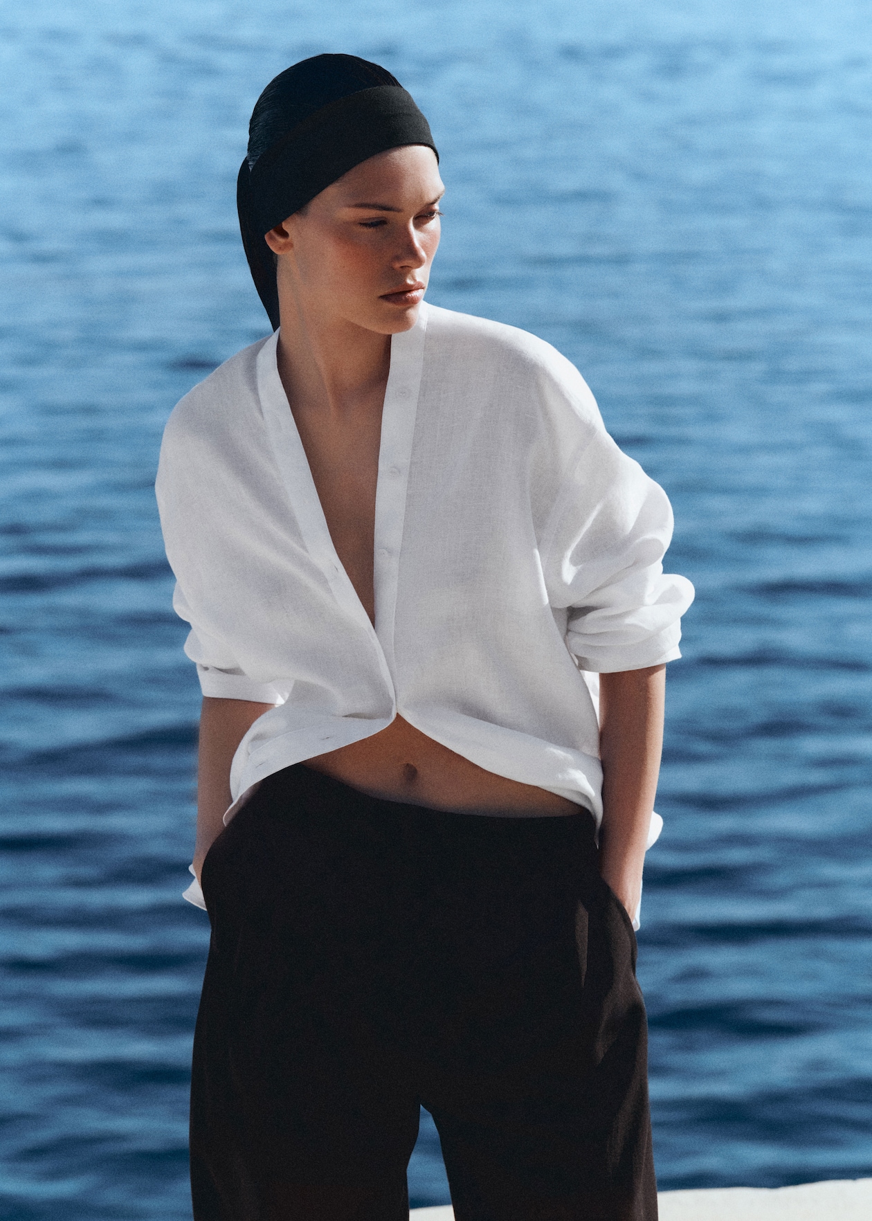 Person wearing a white shirt and black pants stands by a body of water, hands in pockets, with a calm expression and headscarf.