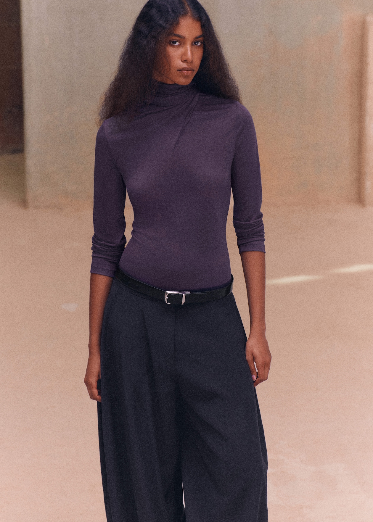 A woman with long brown hair wearing a purple turtleneck and black wide-leg pants stands indoors against a neutral background.