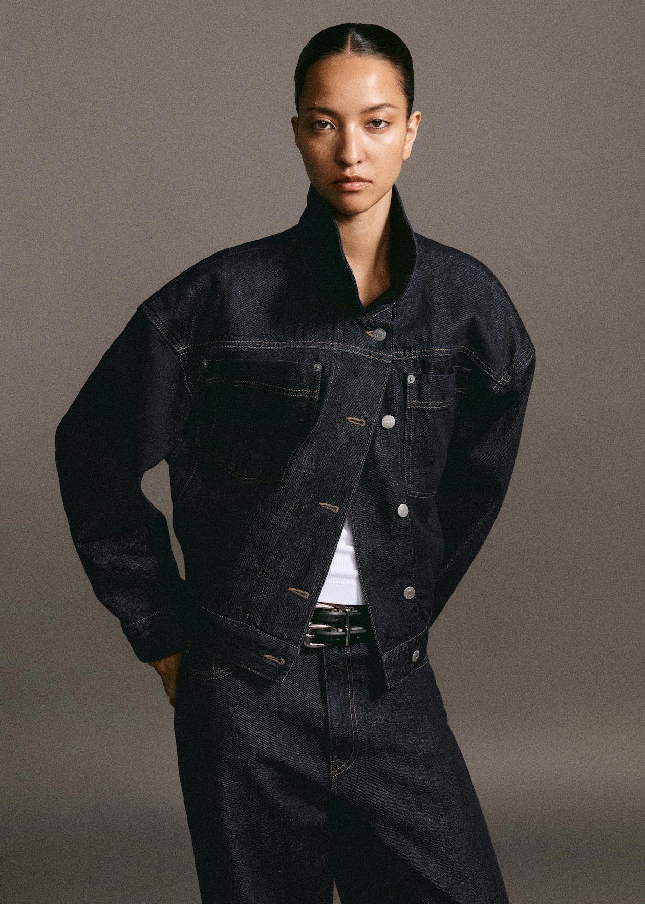 Person wearing a dark denim jacket and jeans with a white shirt, standing against a neutral background, looking directly at the camera.