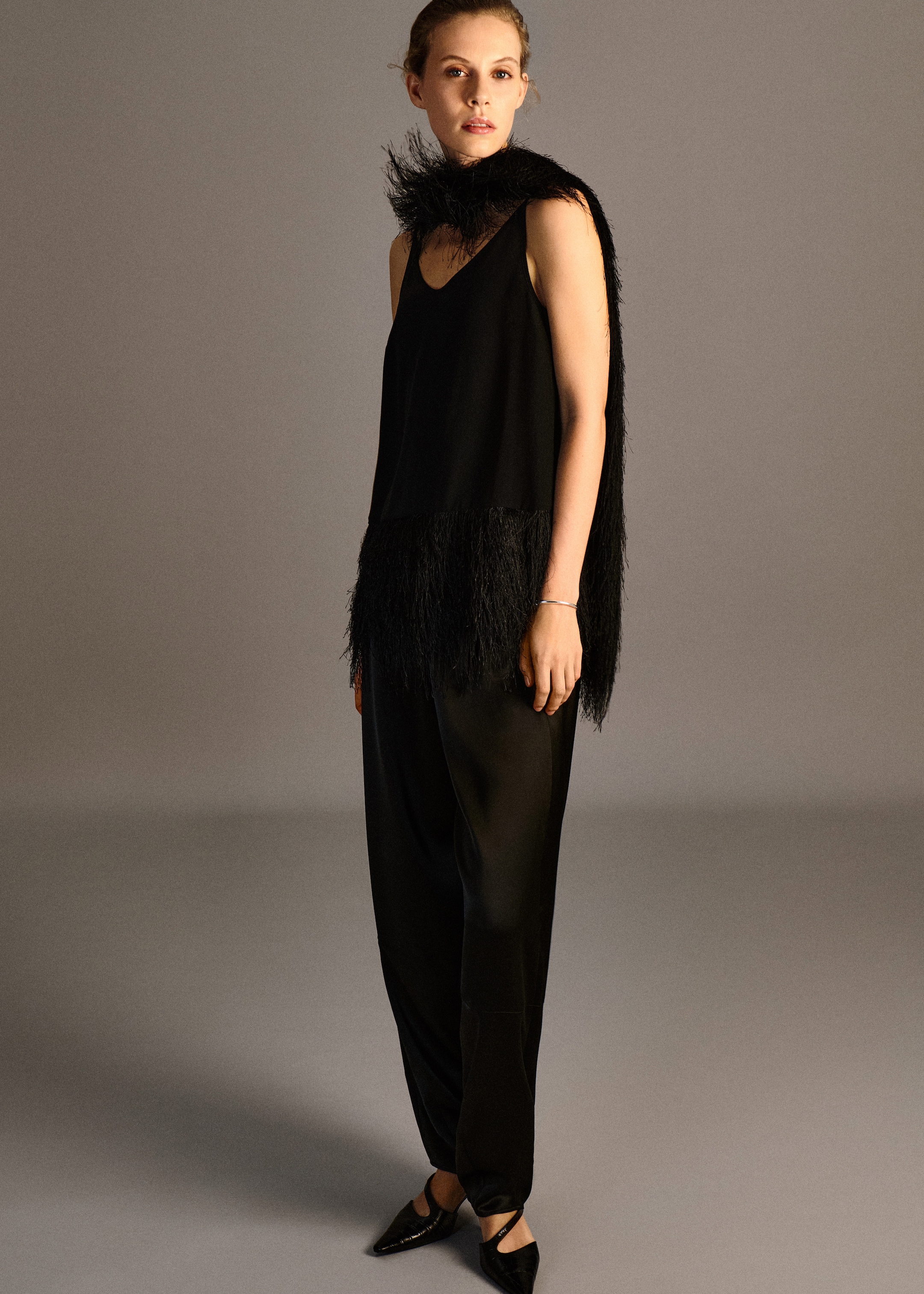 A woman in an all-black outfit with a feathered shawl and pointed shoes, standing against a plain background.
