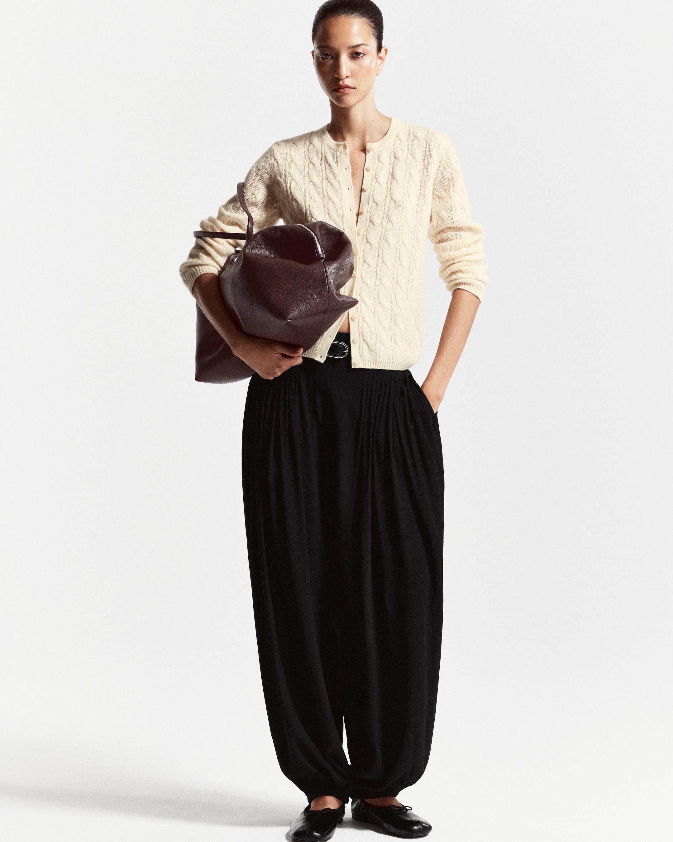 A person wearing a cream cable-knit cardigan and black wide pants holds a large maroon bag, standing against a plain background.