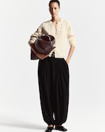 Placeholder for: A person wearing a cream cable-knit cardigan and black wide pants holds a large maroon bag, standing against a plain background.