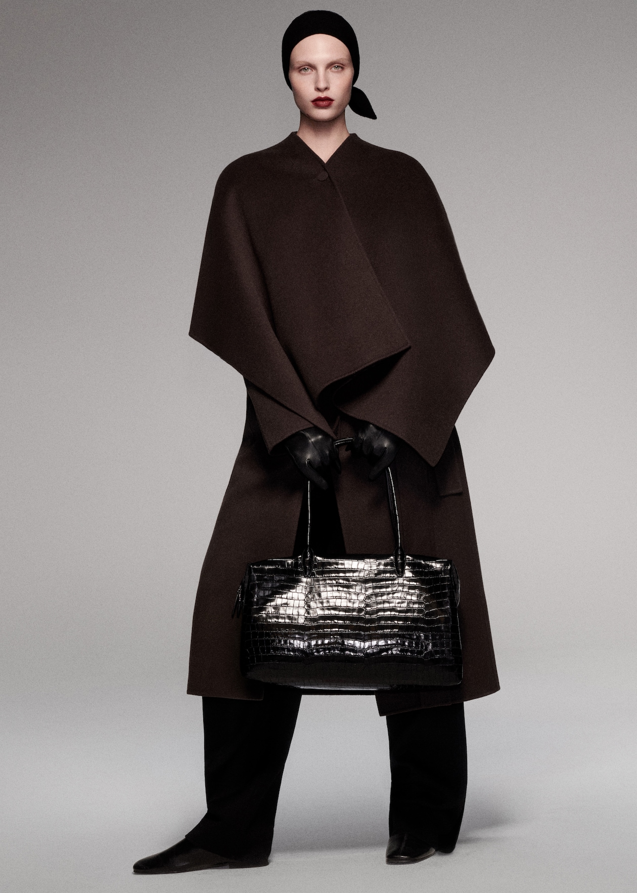 A model wearing a dark, elegant cape, black headscarf, gloves, and holding a large black crocodile-pattern handbag in a minimalist setting.