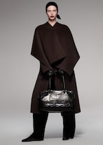 Placeholder for: A model wearing a dark, elegant cape, black headscarf, gloves, and holding a large black crocodile-pattern handbag in a minimalist setting.