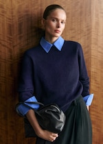 Placeholder for: A person stands against a wooden wall, wearing a navy sweater over a blue shirt, holding a black handbag, and looking directly at the camera.