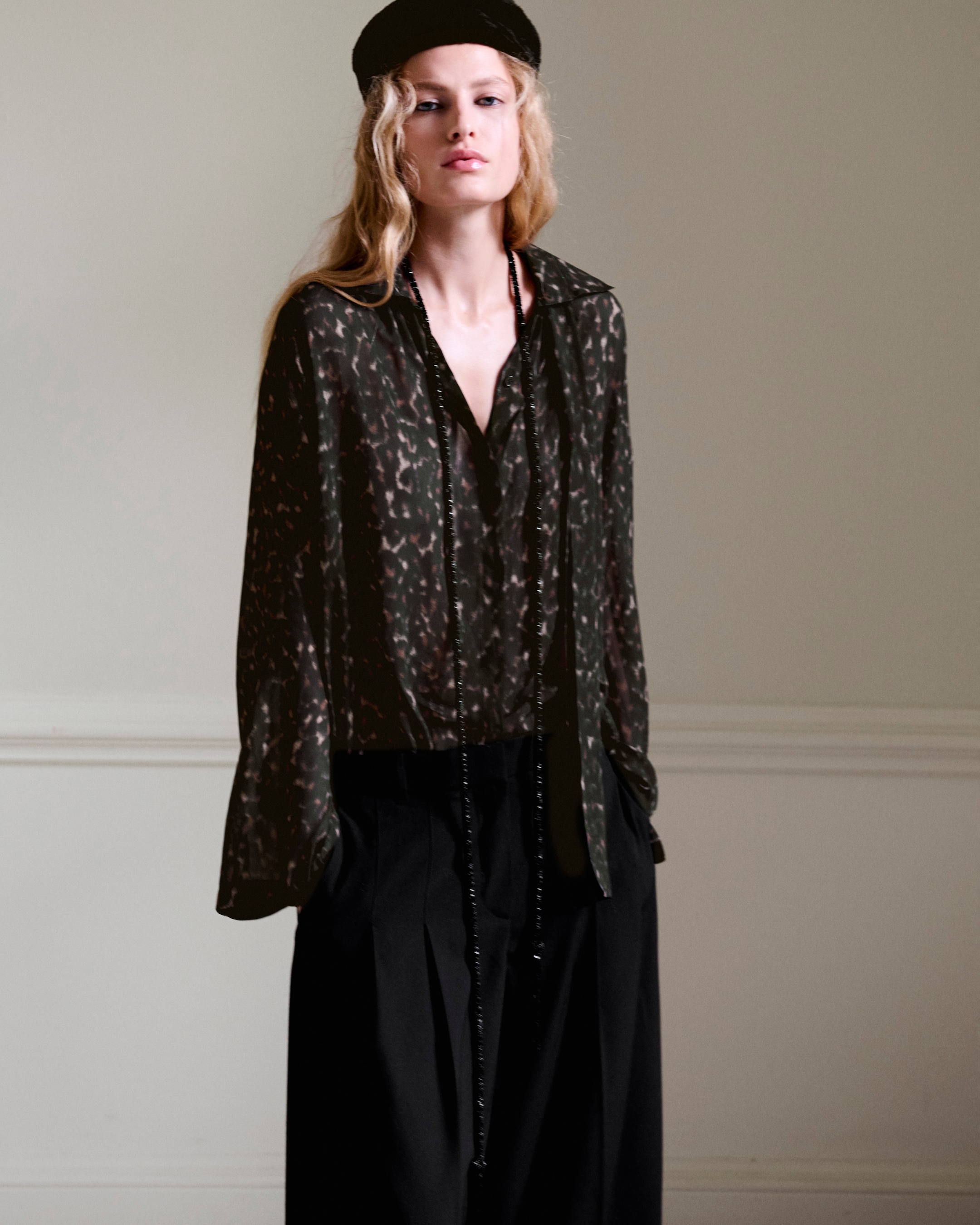 A person wearing a dark patterned blouse, long black pants, and a black beret stands against a neutral background.