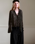 Placeholder for: A person wearing a dark patterned blouse, long black pants, and a black beret stands against a neutral background.