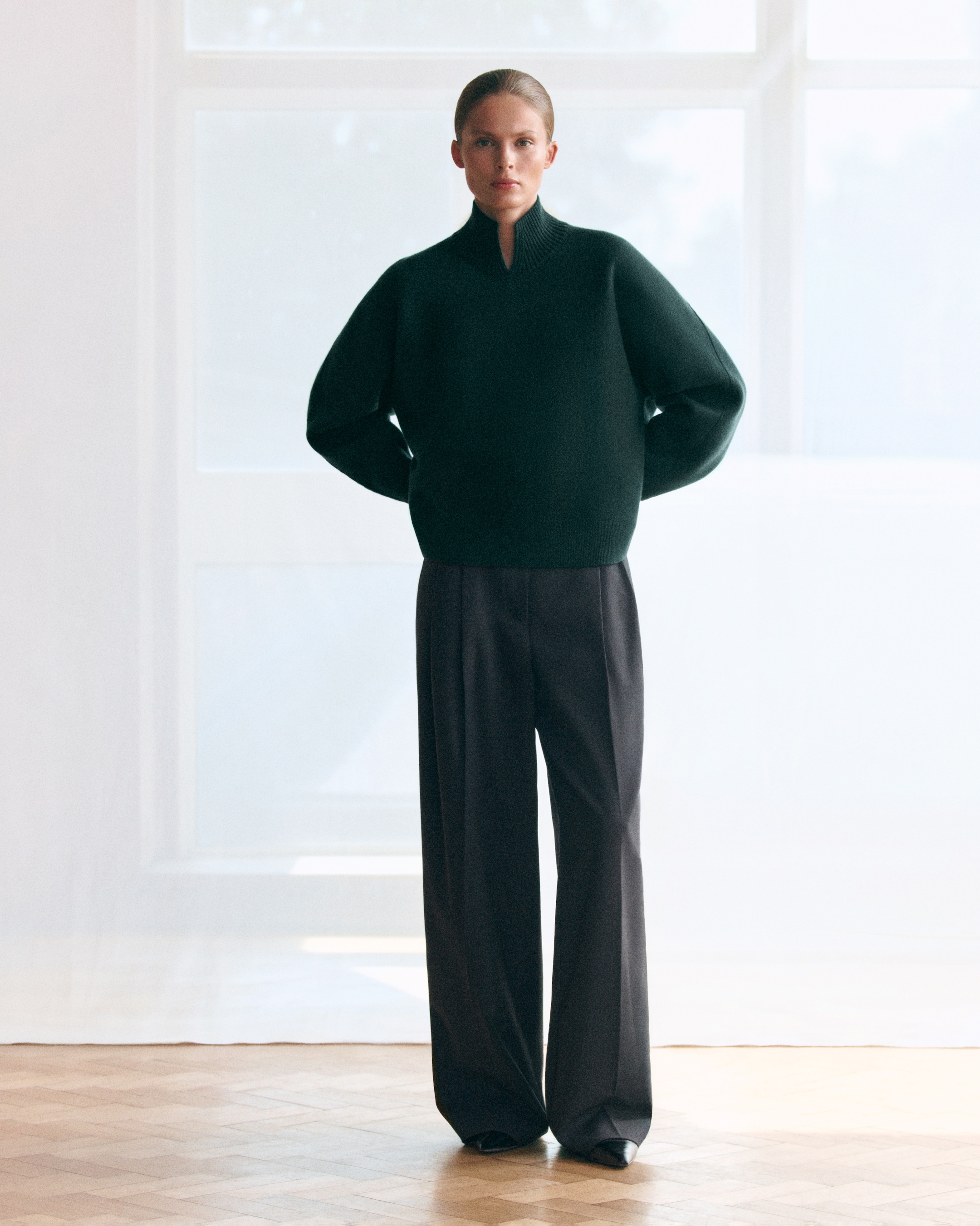 Person standing indoors wearing a dark oversized sweater with a high collar and wide-leg trousers, in front of large, softly lit windows.