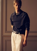Placeholder for: A person with short hair wearing a dark long-sleeve shirt and white pants, standing in a dimly lit room with paneled walls.
