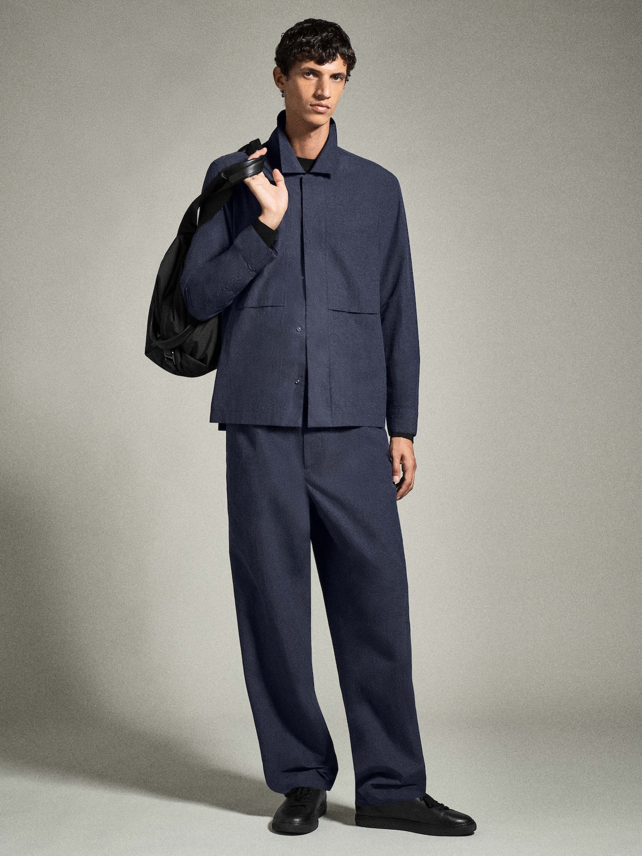 A person wearing a blue matching jacket and trousers, holding a black backpack over one shoulder, standing against a plain background.