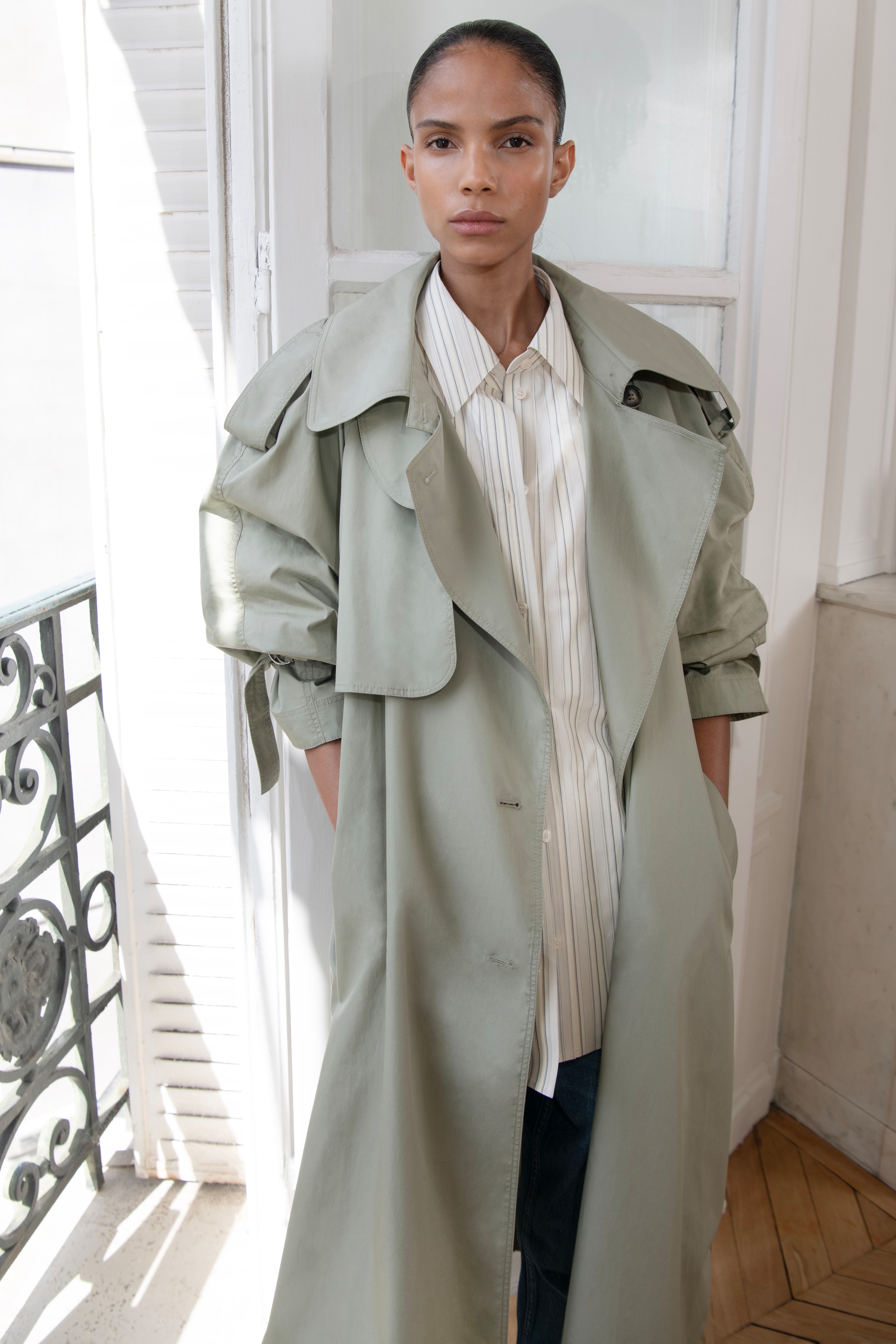 Person in a light green trench coat over a striped shirt, standing in a bright room with hands in pockets, looking at the camera.