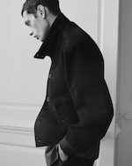 Placeholder for: Black and white profile of a person wearing a dark jacket with a high collar, standing against a light background with hands in pockets.