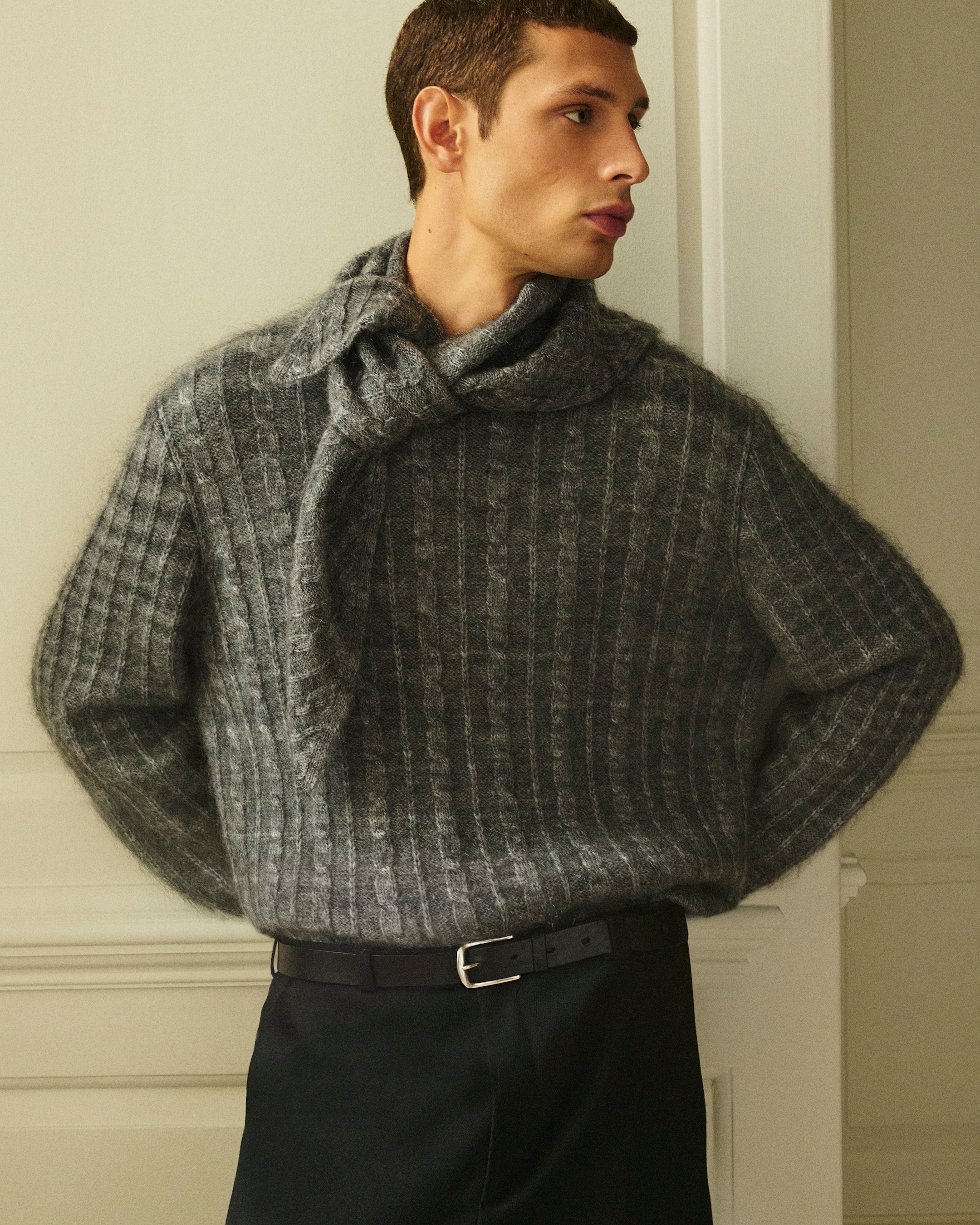 A person wearing a textured gray sweater with a tied scarf detail stands against a light wall, looking to the side.