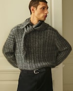 Placeholder for: A person wearing a textured gray sweater with a tied scarf detail stands against a light wall, looking to the side.