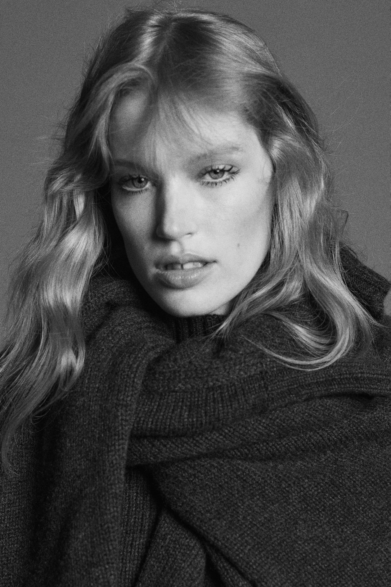 Black and white portrait of a person with long blonde hair, wearing a dark sweater, looking directly at the camera with a soft, serious expression.