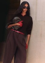 Placeholder for: A stylish woman wearing sunglasses, a dark top, and wide-leg trousers, holds a bag. She accessorizes with a red belt and gloves.