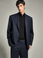 Placeholder for: A man in a dark navy suit with a black turtleneck stands with his hands in his pockets against a light gray background.