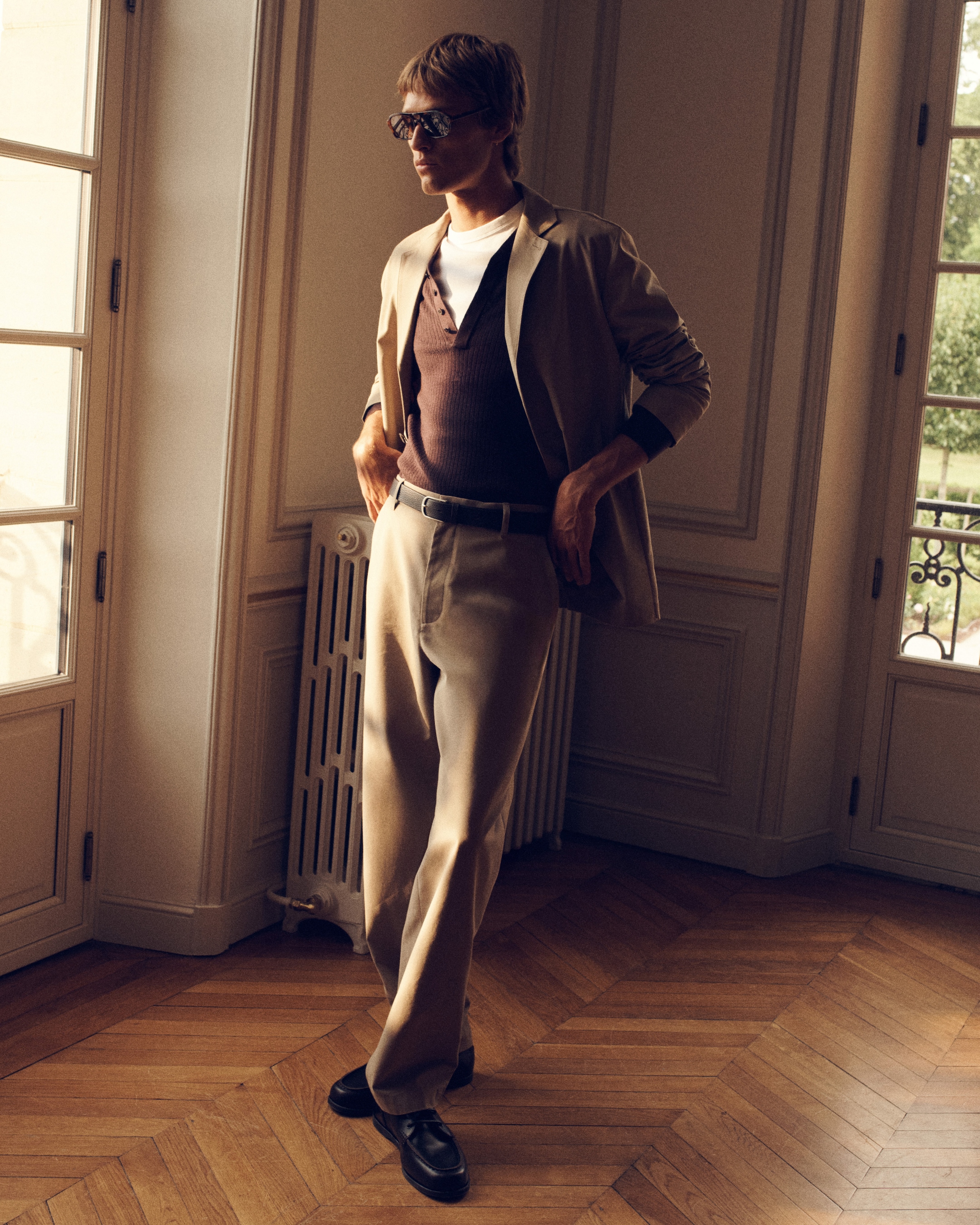 A person in sunglasses stands near a window, wearing a beige suit over a brown shirt and white undershirt, in a sunlit room with wooden flooring.