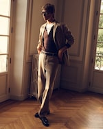 Placeholder for: A person in sunglasses stands near a window, wearing a beige suit over a brown shirt and white undershirt, in a sunlit room with wooden flooring.