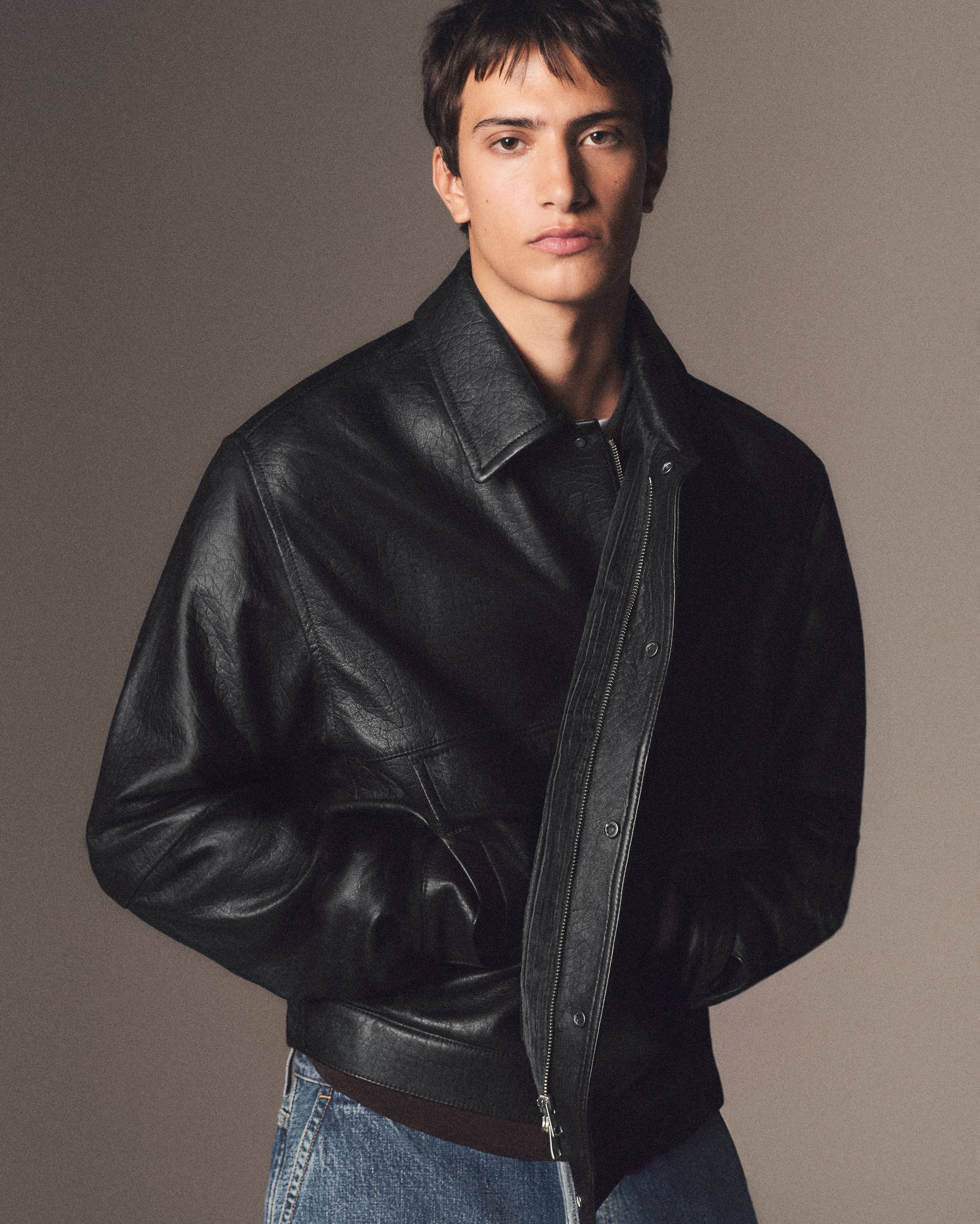 Person wearing a black leather jacket and jeans stands against a neutral background.