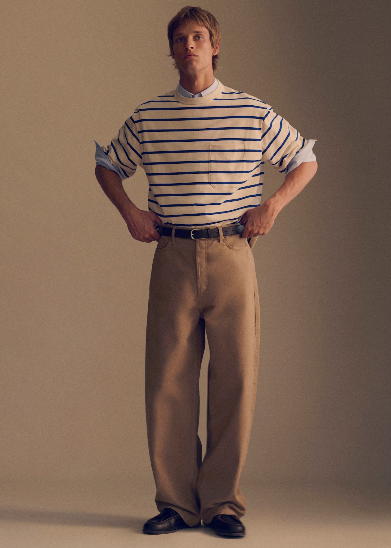 A person stands confidently wearing a striped shirt over a collared shirt, loose-fitting khaki pants, and black shoes, hands on hips.