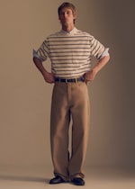 Placeholder for: A person stands confidently wearing a striped shirt over a collared shirt, loose-fitting khaki pants, and black shoes, hands on hips.