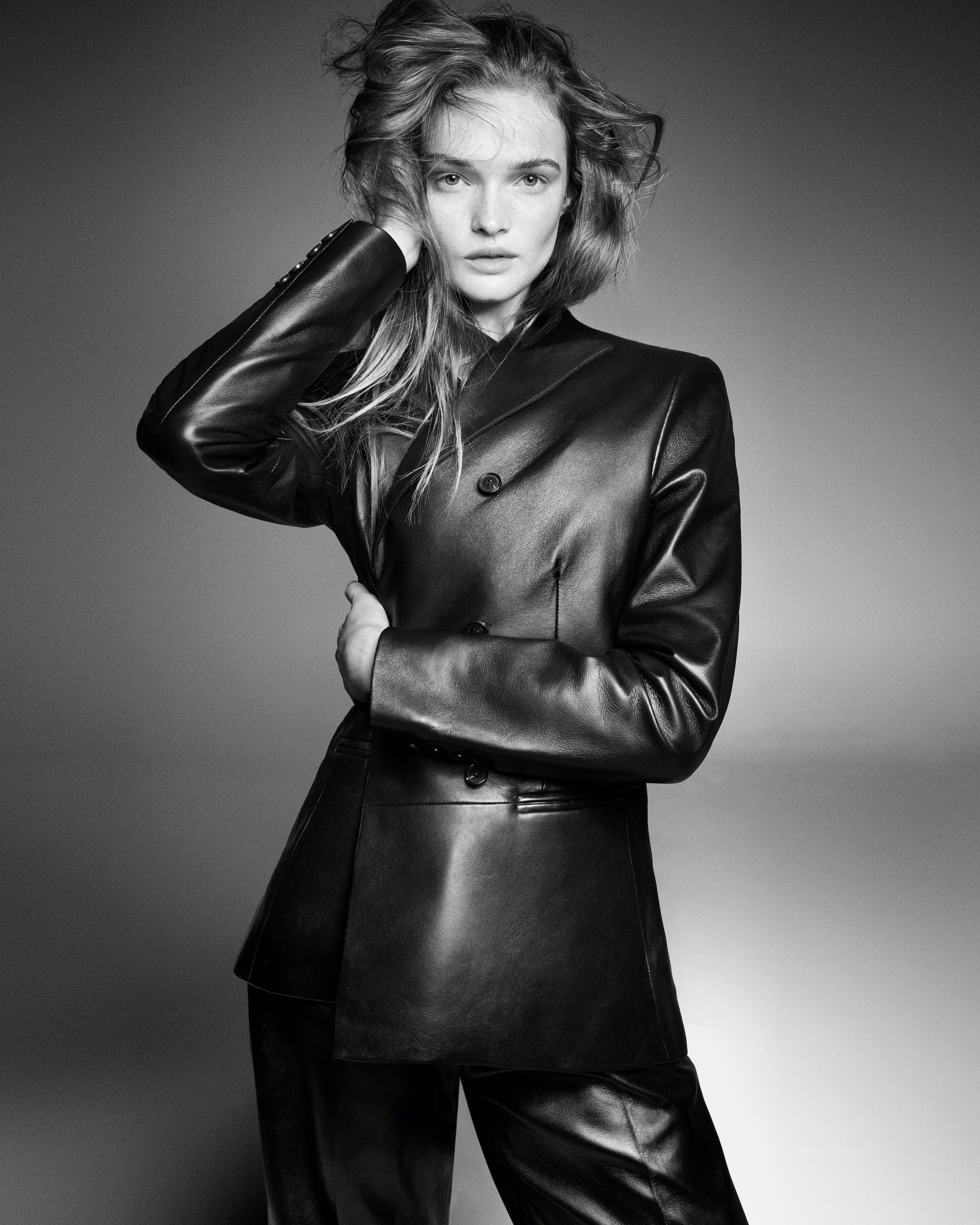 Model wearing a sleek, double-breasted leather suit poses with one hand touching her hair in a dramatic black and white portrait.