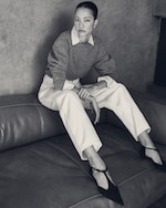 Placeholder for: Woman sitting on a leather couch wearing a sweater over a striped shirt, white pants, and black heels, looking at the camera. Black and white image.