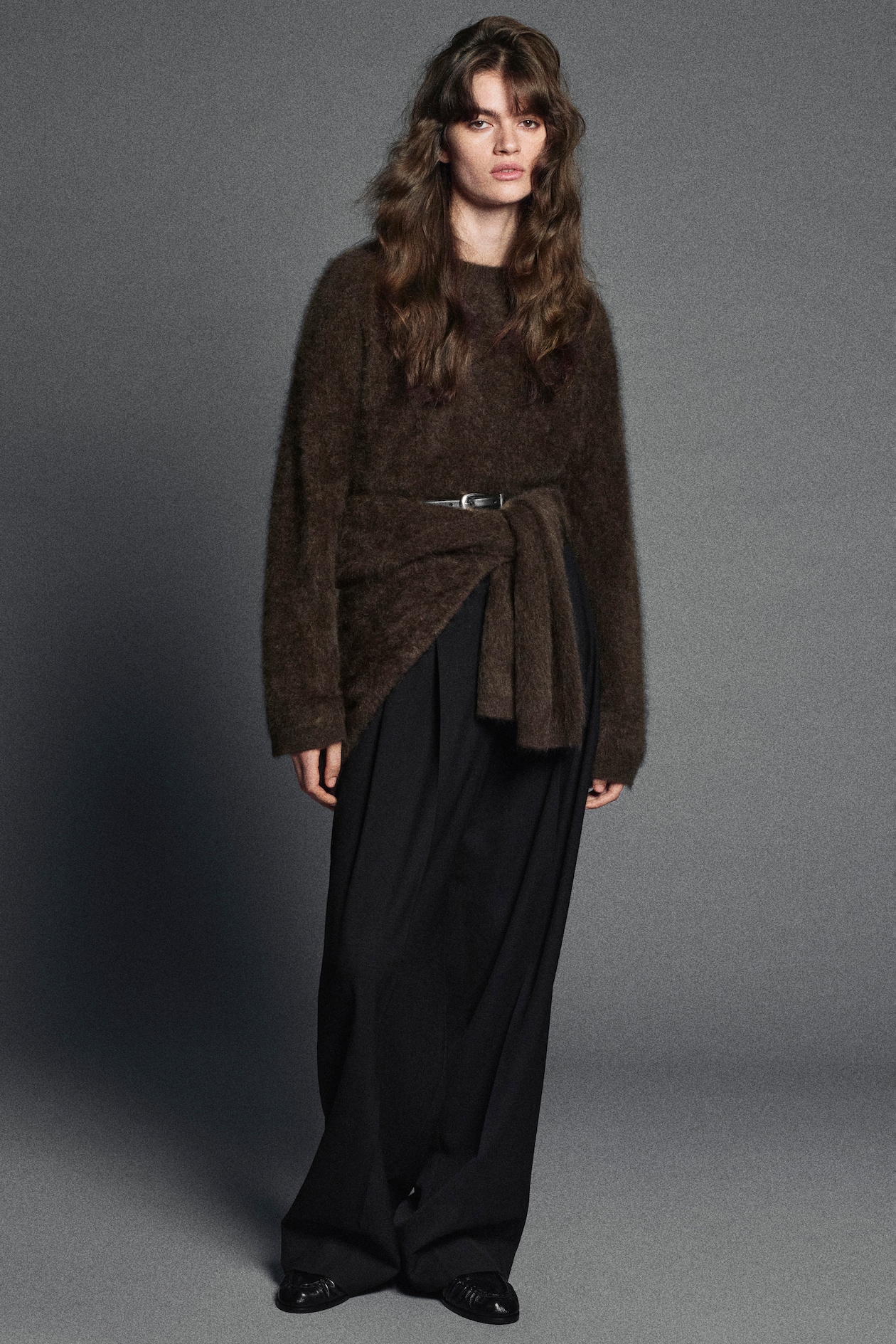 Woman wearing a brown fluffy sweater with a belt, black wide-leg pants, and black shoes, posing against a gray background.