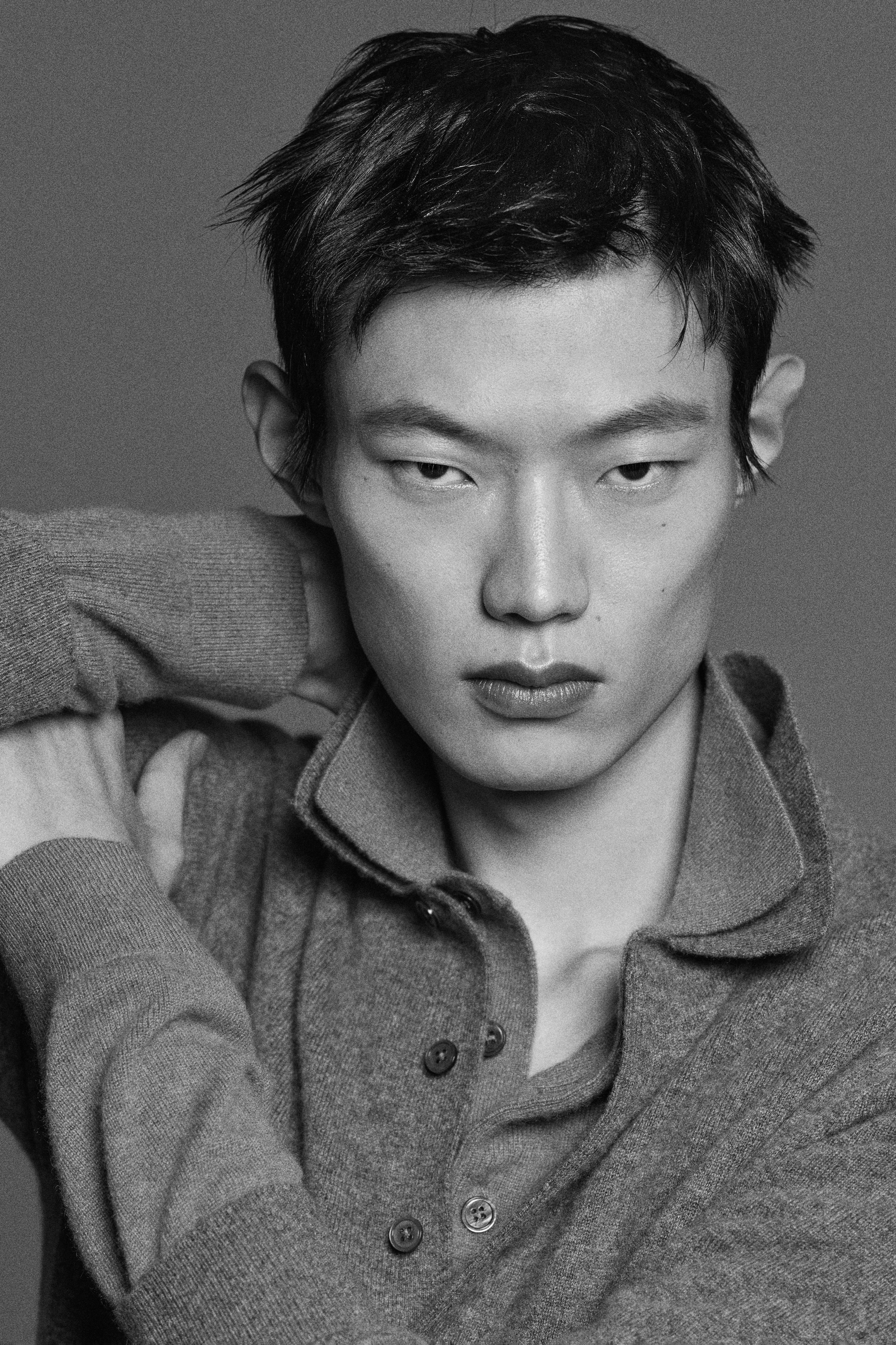 Black and white portrait of a person with short hair, looking directly at the camera, wearing a buttoned-up collared sweater with hands behind the head.