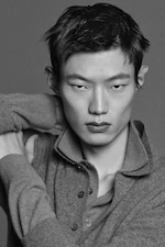 Placeholder for: Black and white portrait of a person with short hair, looking directly at the camera, wearing a buttoned-up collared sweater with hands behind the head.