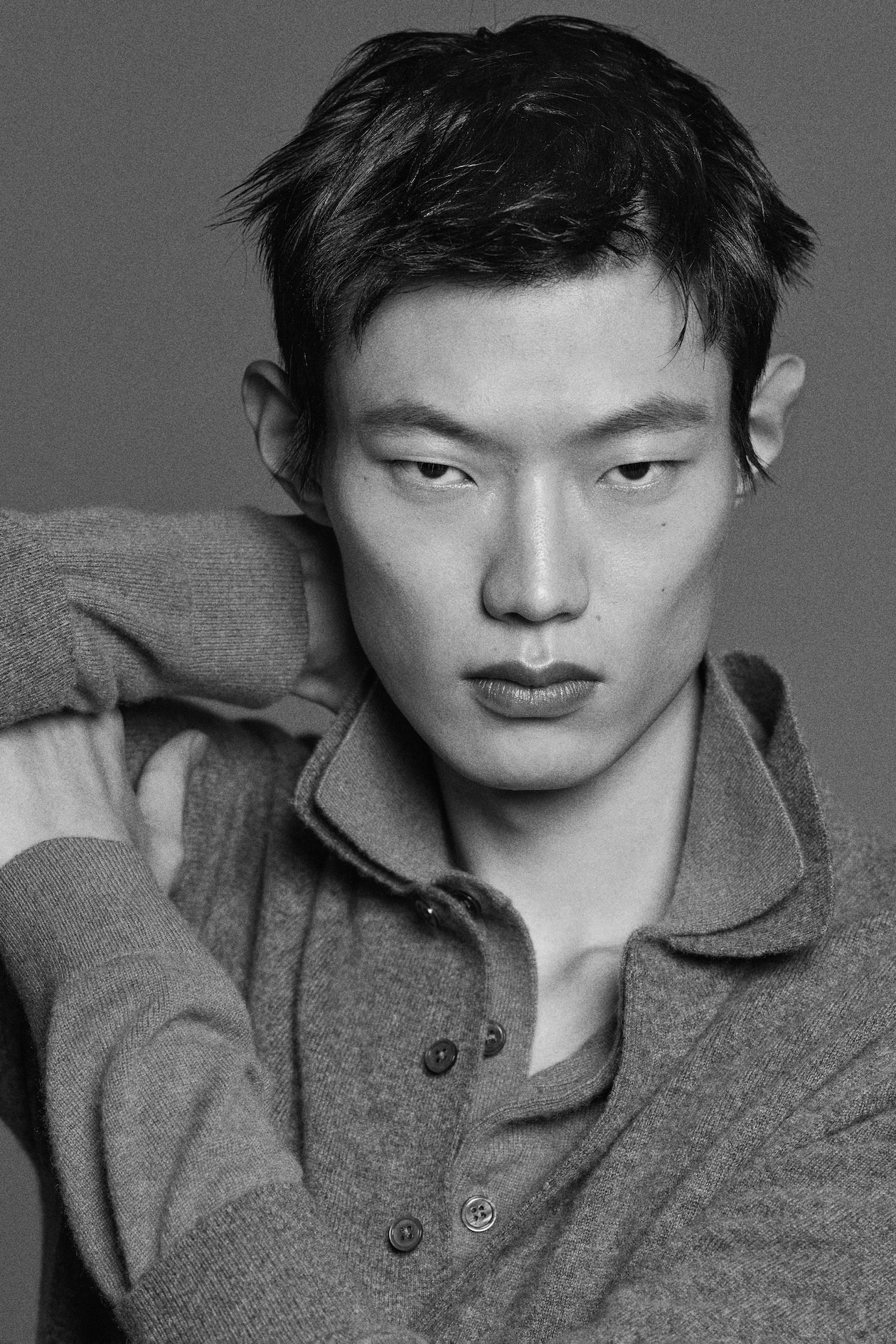 Black and white close-up of a person with short hair, wearing a buttoned sweater, posing with arms raised behind their head, looking directly at the camera.