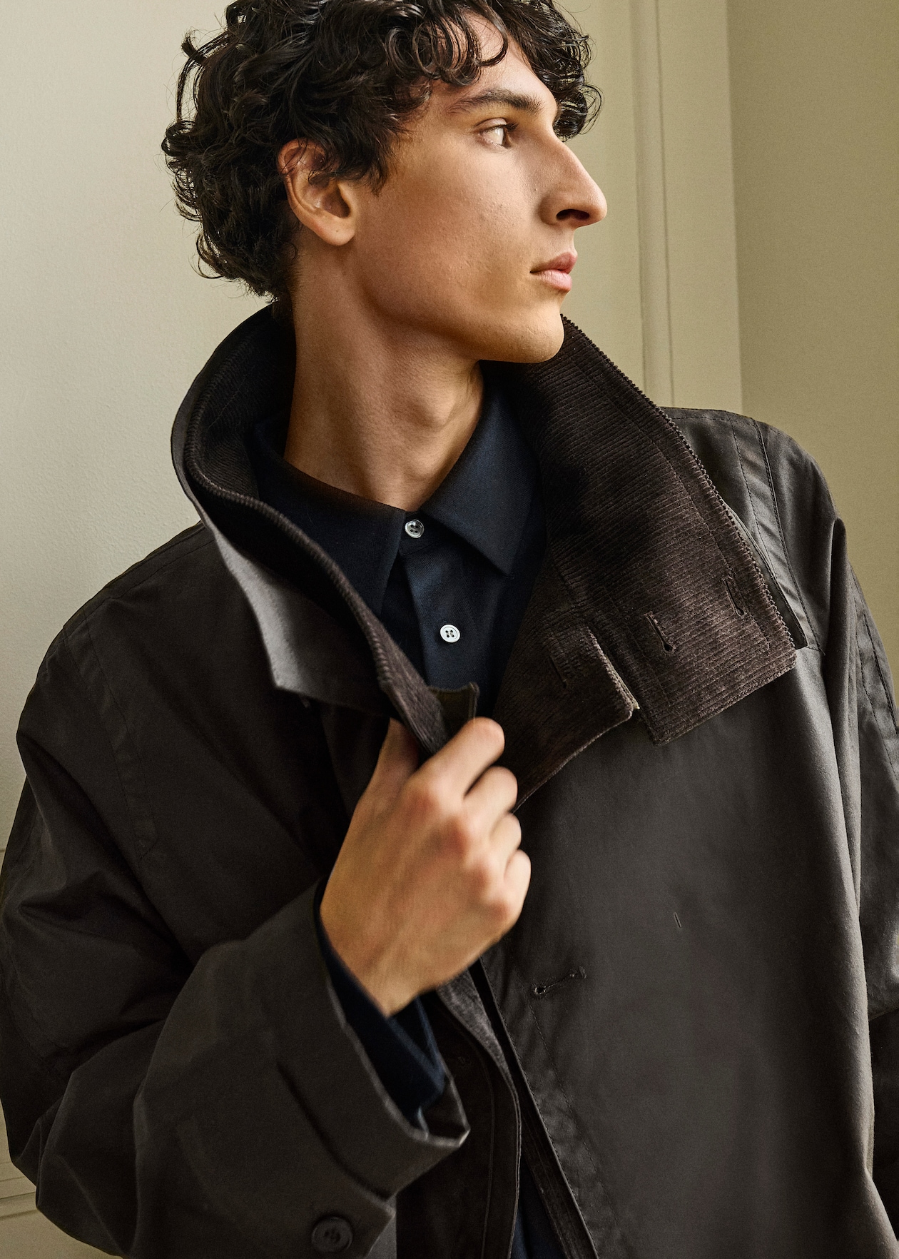 A person with curly hair wearing a dark jacket and shirt looks to the side, adjusting their collar.