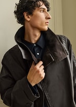 Placeholder for: A person with curly hair wearing a dark jacket and shirt looks to the side, adjusting their collar.