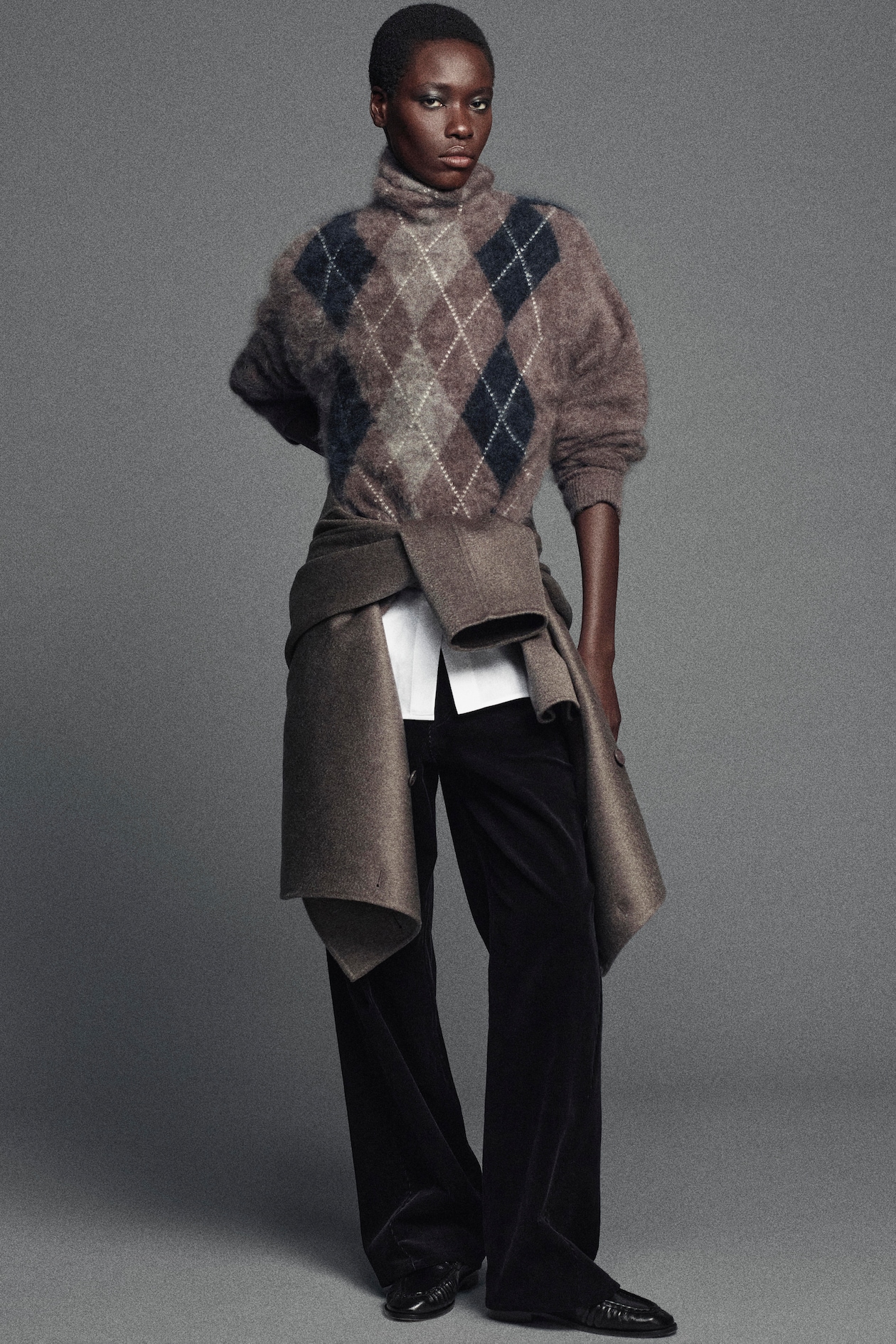 Person wearing a brown argyle sweater, dark pants, with a coat tied around their waist, against a gray background.