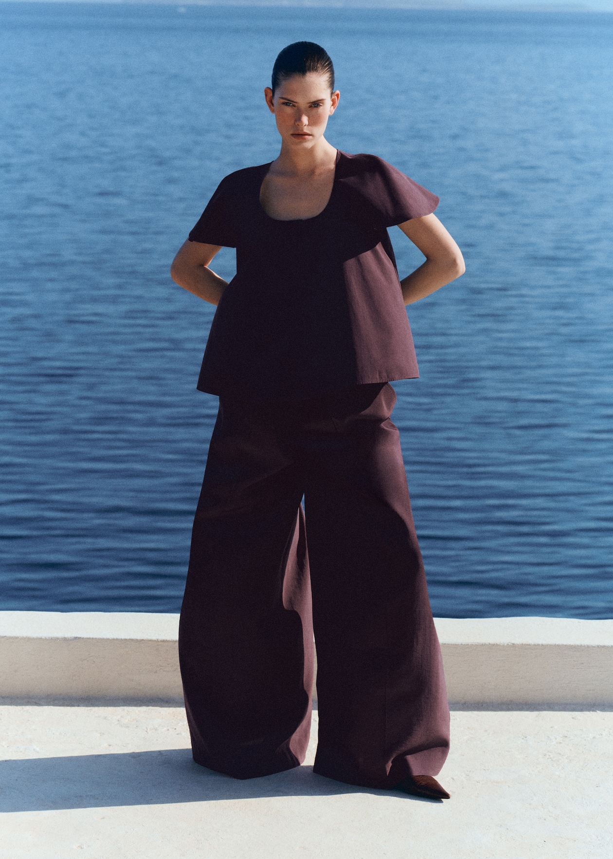 Person in a burgundy outfit stands confidently by the sea, with a clear blue sky and water as the backdrop.