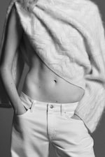 Placeholder for: A black and white photo of a person wearing a soft knit sweater draped across the chest and white jeans, hands in pockets, highlighting their midriff.