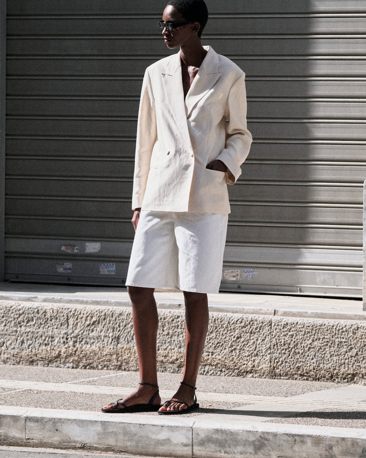 Person in a beige blazer and white shorts stands on a sunlit pavement, wearing sunglasses and sandals, with a shuttered backdrop.
