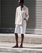 Placeholder for: Person in a beige blazer and white shorts stands on a sunlit pavement, wearing sunglasses and sandals, with a shuttered backdrop.