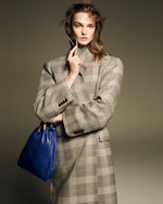 Placeholder for: Model in a plaid coat holding a blue bag with one hand, the other hand touching her face, set against a neutral background.
